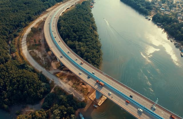 Aerial shot of vehicles on a curved highway bridge crossing a tranquil river surrounded by lush greenery.