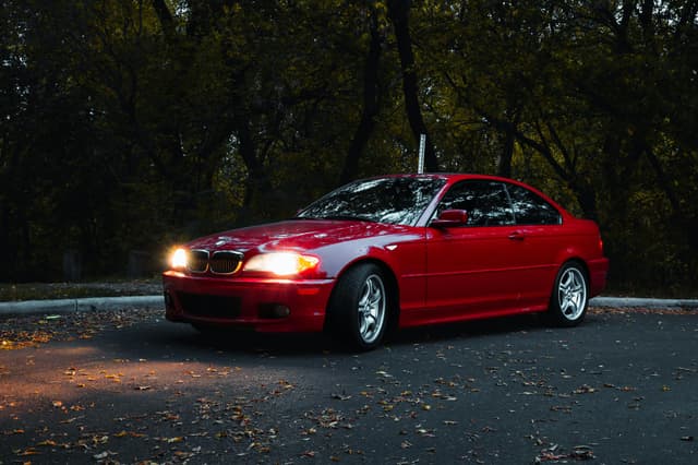 A red car parked outdoors with headlights on, surrounded by autumn trees in Edmonton, Canada.