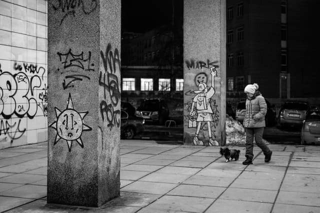 A woman walks her dog past graffiti in a city at night. Urban life captured in monochrome.
