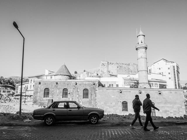 Monochrome capture of a street scene featuring an old car and walking individuals near Ottoman architecture in Bitlis, Türkiye.