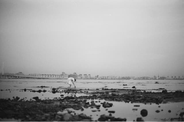 Grayscale photo of a person by the rocky shore with city skyline in Novorossiysk.