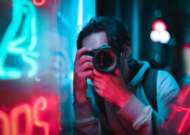 A young man photographing neon lights in an urban setting, showcasing vibrant colors and creativity.