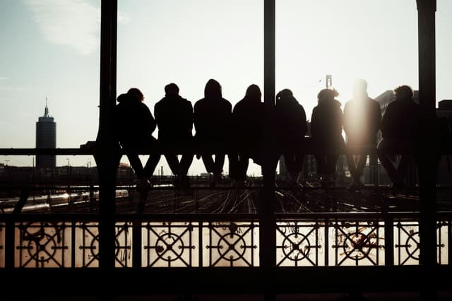 Group of seven people sitting on a bridge railing, silhouetted against a sunset sky in an urban setting.