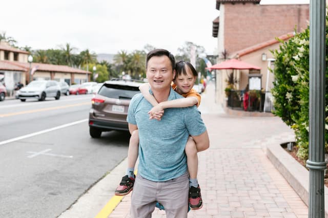 A joyful Asian father giving his child a piggyback ride on a city street.
